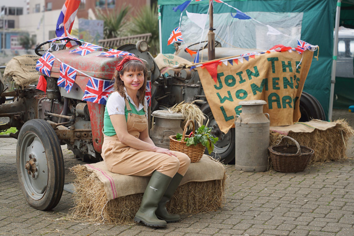Land Girl sits on hay bail in front of tractors