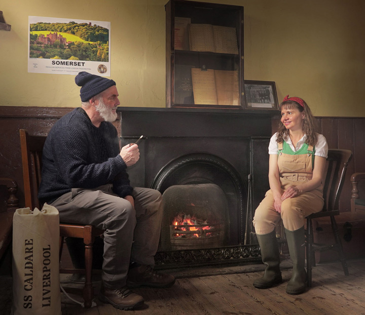 Sailor Tim and land girl Asia sit next to fire in train station wating room